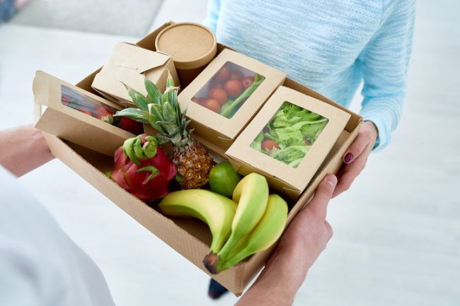 En person tar emot en matkasse med färsk frukt och färdiga sallader i pappkartonger.